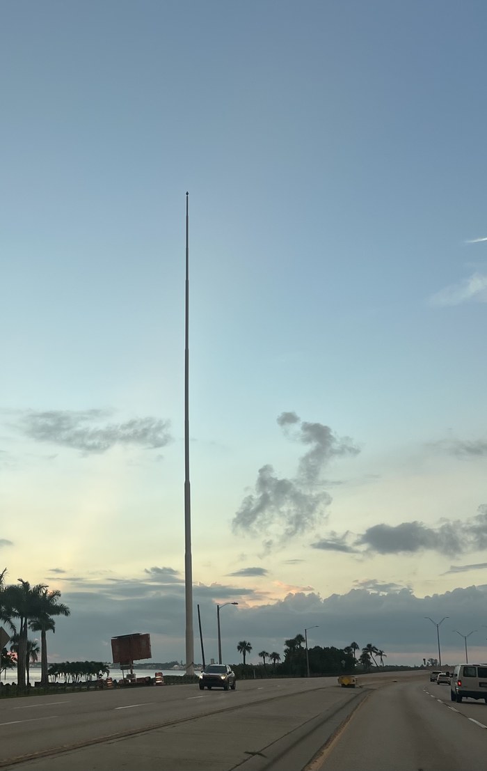 Cape Coral's 250-foot flagpole standing bare with no flag against a sunset sky, photographed from the road near the Cape Coral Bridge